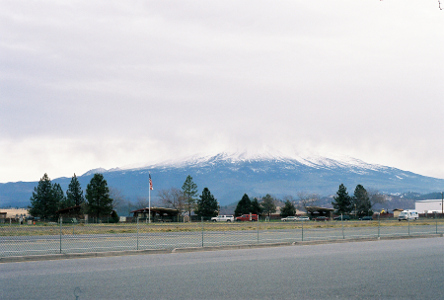 [Hardly any snow is visible on the mountain because the top part of the mountain is completely covered in clouds and not visible. There is some snow at the lower level which is visible before tapering downward to green growth. There is no precipitation on the roadway even though the sky is completely overcast.]